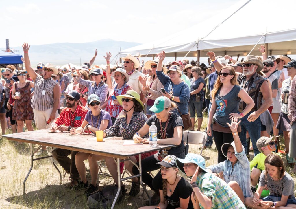 Outdoor festival crowd gathered beside a tent, with people raising their hands and sitting at a table.